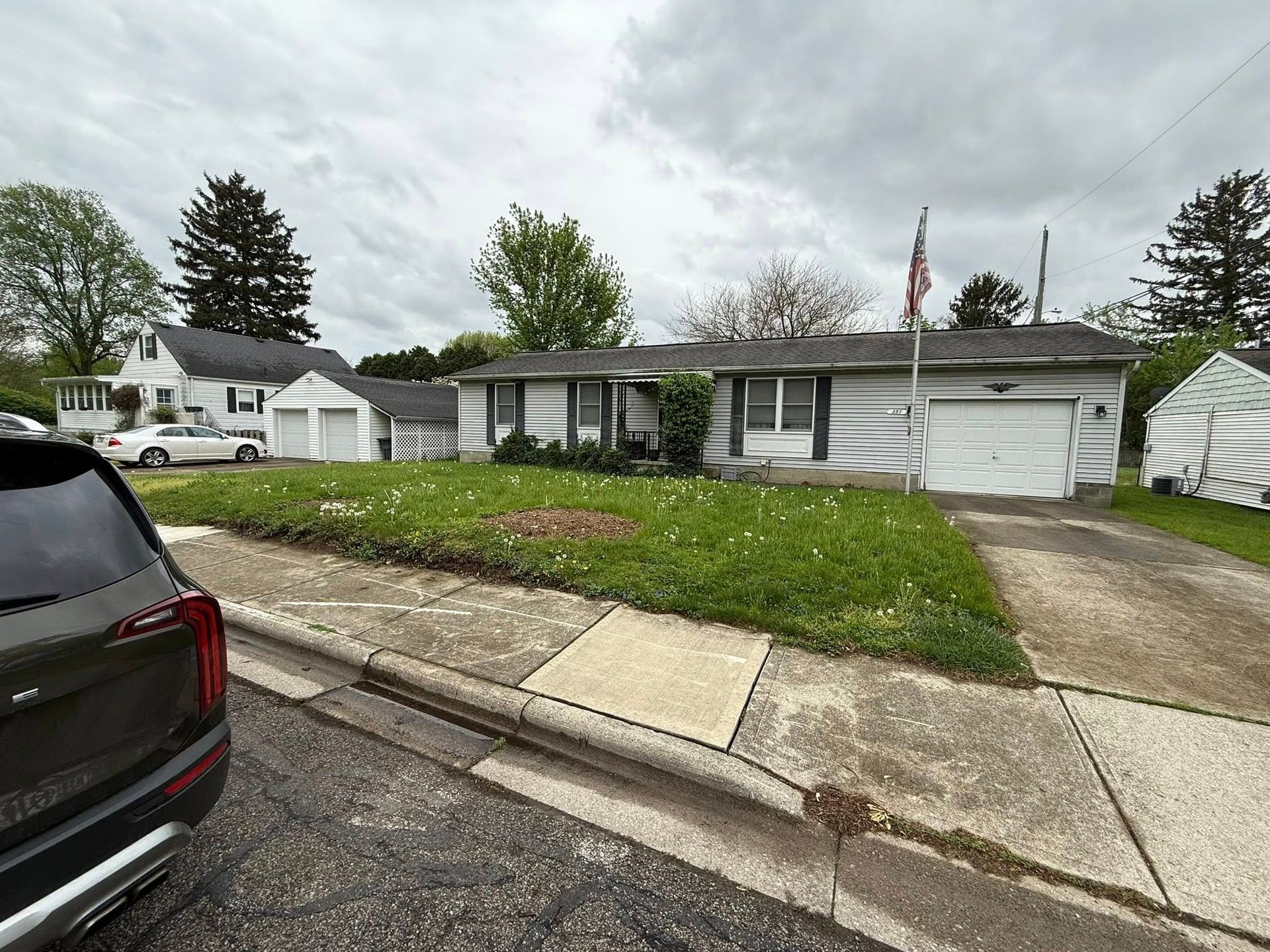 A ranch-style house with overgrown lawn, attached garage, and a flag, under a cloudy sky.