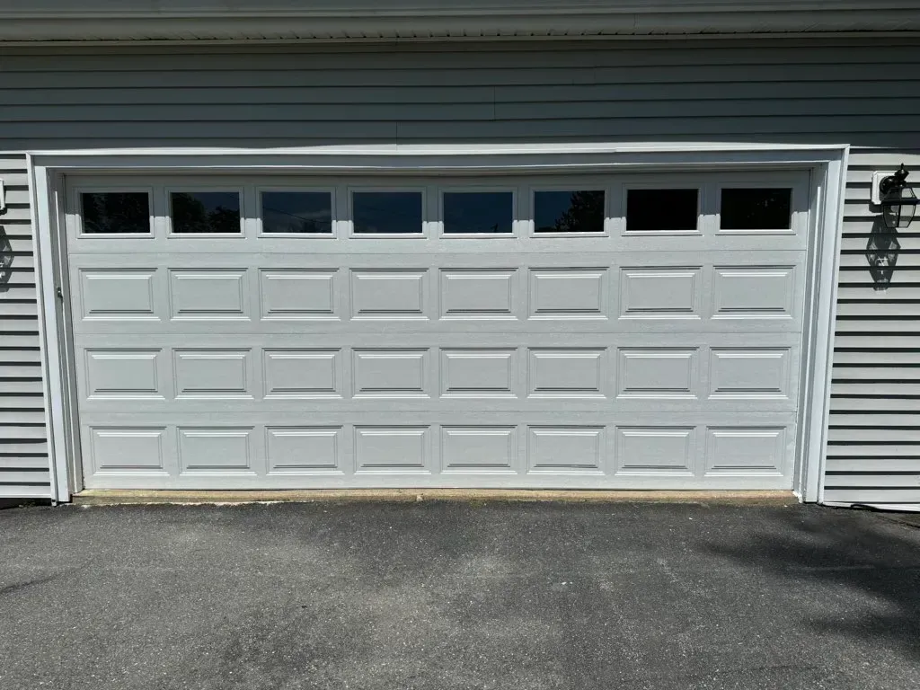 White garage door with windows, set in a light blue house, on an asphalt driveway.