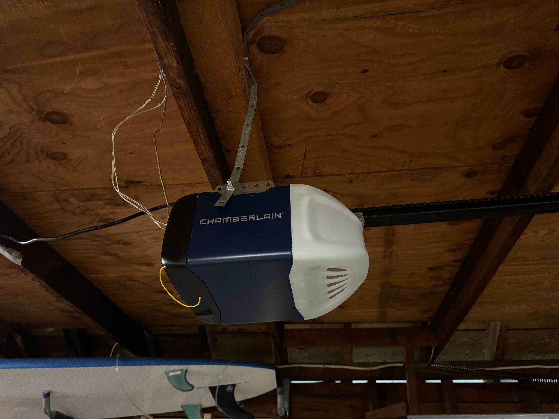 Garage door opener mounted on a wood ceiling. Blue and white opener with wires hanging.