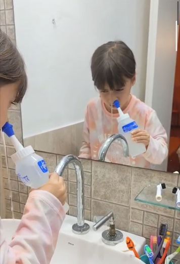 Una persona se cepilla los dientes en el lavabo del baño, reflejada en el espejo, con un tubo de pasta de dientes y un cepillo de dientes.