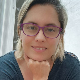 Una persona con gafas moradas sonríe en un retrato de primer plano, apoyando la barbilla en la mano.