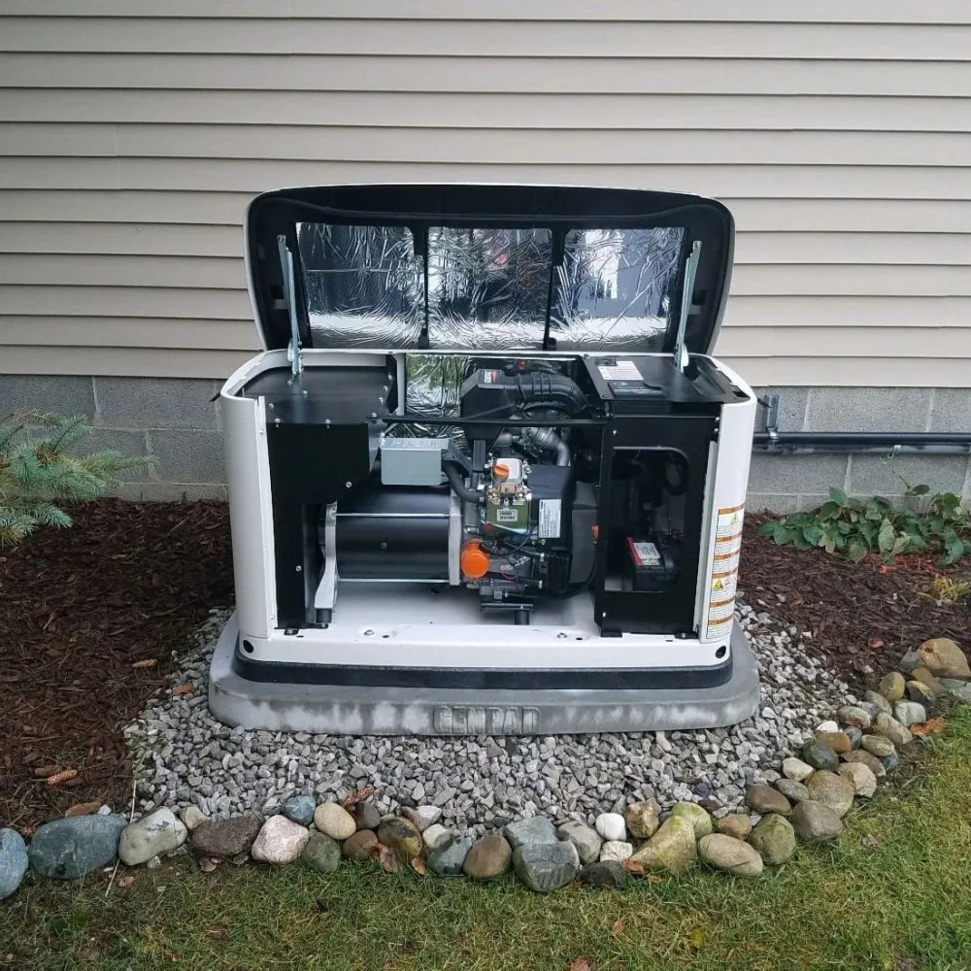 Backup generator with open panels, tools visible, mounted on gravel base beside house.