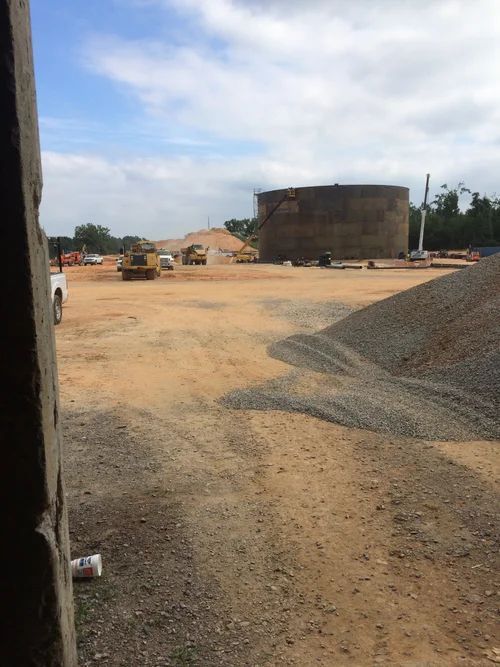 Construction site with a large concrete cylinder, heavy machinery, and gravel piles on a dirt lot under a partly cloudy sky.