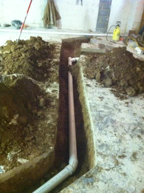 Trench in concrete floor with PVC pipes laid for plumbing; dirt and tools surrounding.