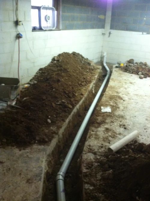 A trench dug for installing a gray pipe, with a pile of dirt inside a room with a window.