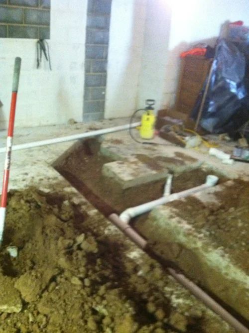 Open trench in a concrete floor, showing plumbing pipes. Construction site, with tools and equipment.