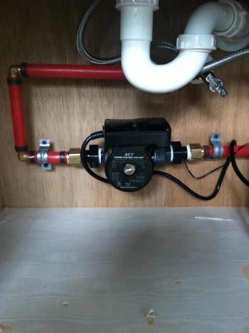 Water recirculating pump installed under a sink, connected to red pipes and white plumbing.