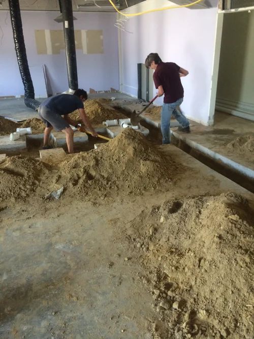 Two people using shovels to move dirt in a room under construction.