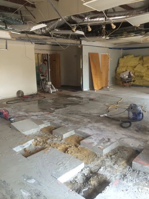 Interior demolition of a room with concrete floor. Worker operates a saw; exposed ceiling, debris.