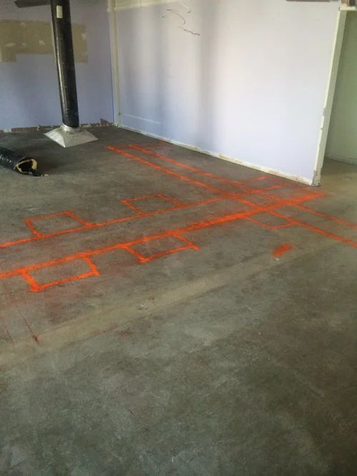 Orange spray-painted outlines on a concrete floor, indicating potential floor plan layout in a room with white walls.