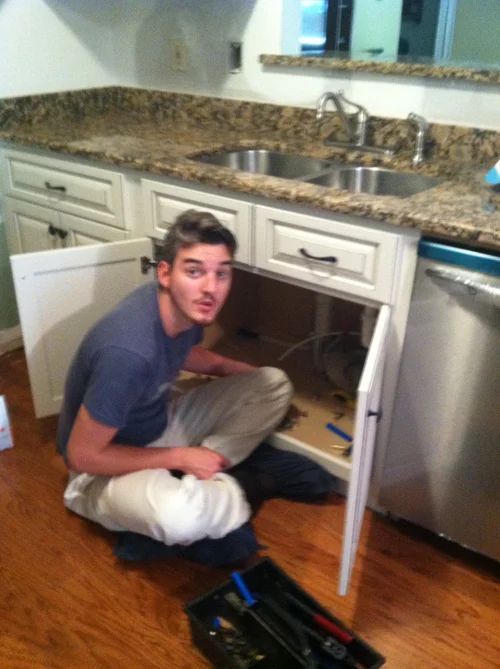 Man crouches in a kitchen cabinet working on plumbing under a sink. He wears a blue shirt and white pants.