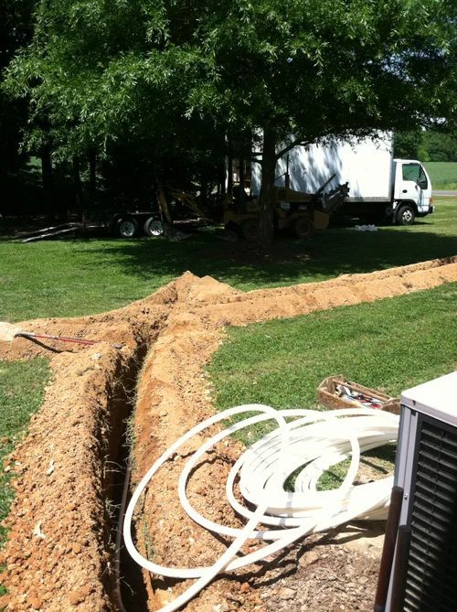 A trench in grass with white tubing next to a white truck. Sunny outdoor setting.
