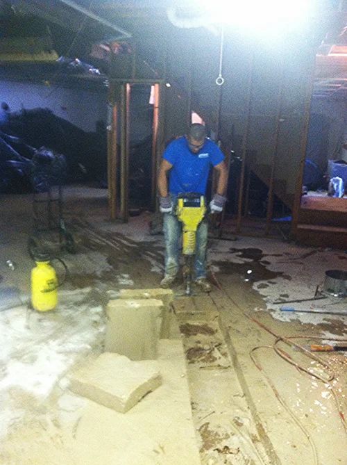 Man in blue shirt using jackhammer to break up concrete in a basement.