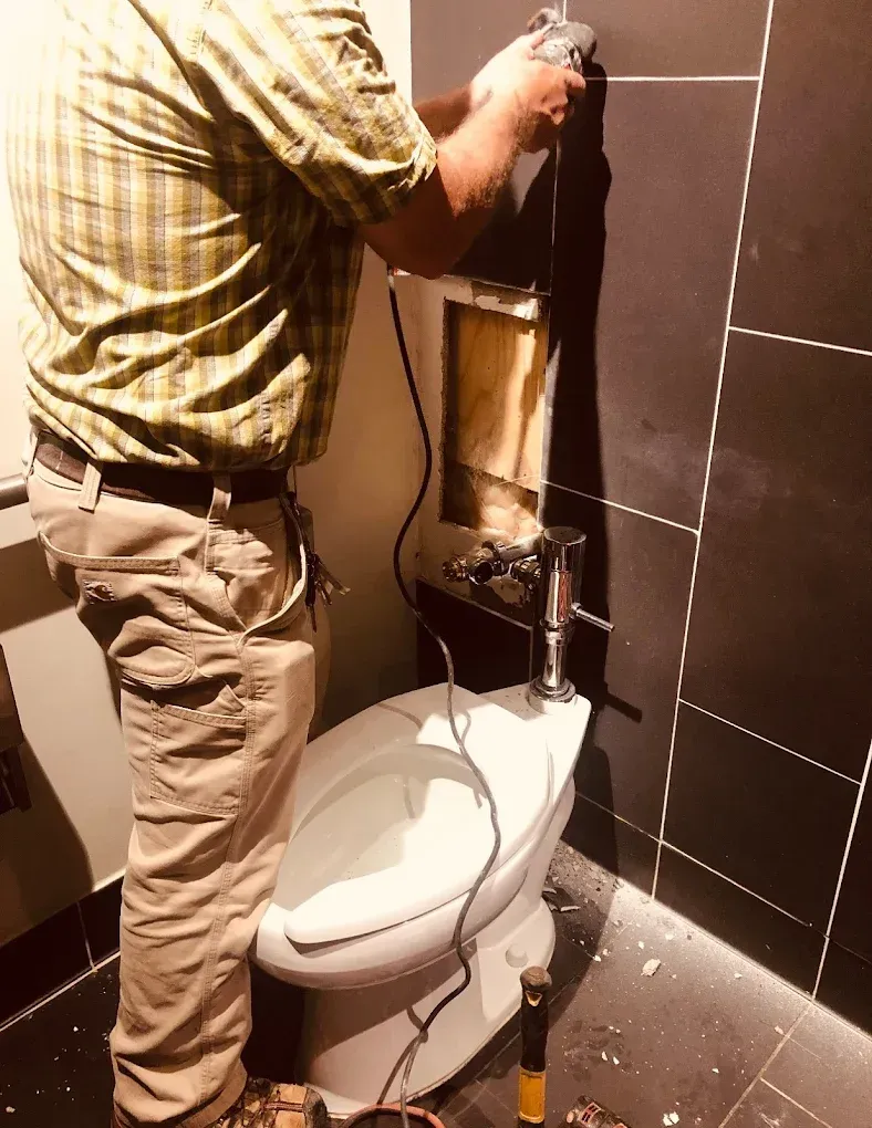 A person repairs bathroom wall behind a toilet, using a power tool, with dark tile surround.