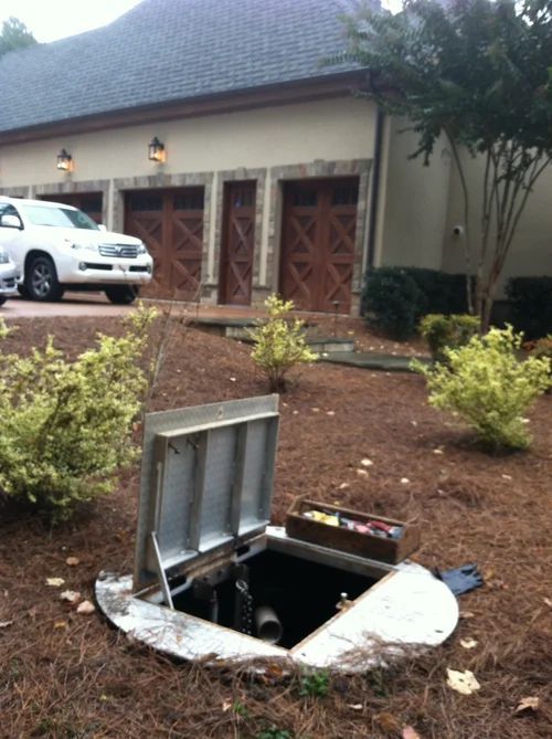 Open utility access hatch in a yard with tools visible. A house and car are in the background.