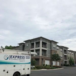 A white Express Plumbing Services truck parked in front of a large apartment building on a cloudy day.