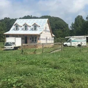 House under construction with trucks, a green field, and trees.