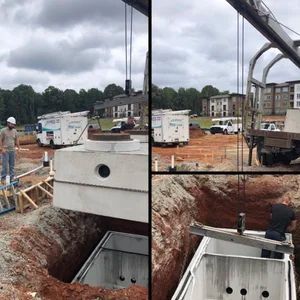 Construction site: crane lowers a concrete structure into an excavated pit. Workers in the background.