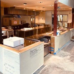 Restaurant kitchen under construction; stainless steel appliances and counters, boxes, drywall, and cement floor.