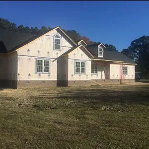 House under construction, beige exterior, windows, black roof, blue sky, grass.