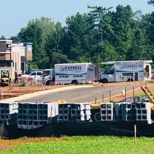 Construction site with Express trucks, blocks, and workers; trees and buildings in background.