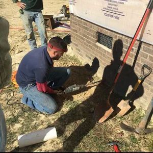 Man kneeling, cutting near a brick wall. His shadow and tools are visible. Outdoors, daytime.