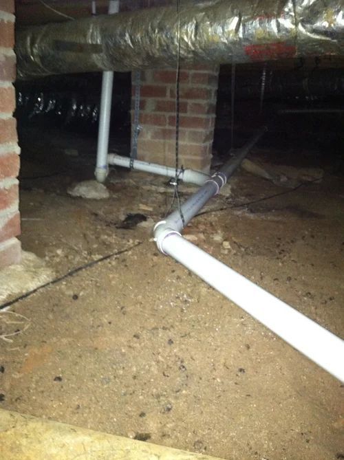 Underneath a house, white PVC pipes run along the dirt floor. Brick supports a duct. Dark, enclosed space.
