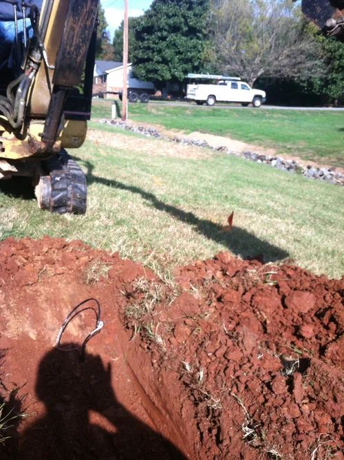 An excavator digging in reddish dirt, shadow of person taking photo, white van in background.