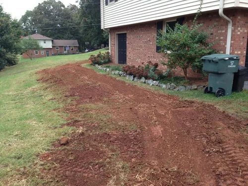 A path of red dirt, likely for a driveway, is being constructed on a grassy hill next to a brick house.