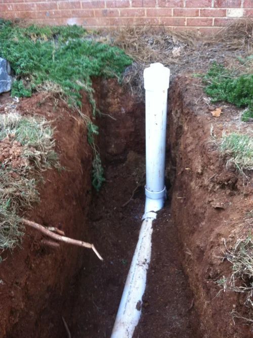 White PVC pipes in a trench, near a brick wall and grass.