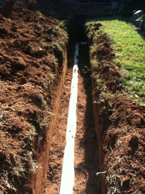 Trench dug in soil, containing white pipe, alongside green grass.
