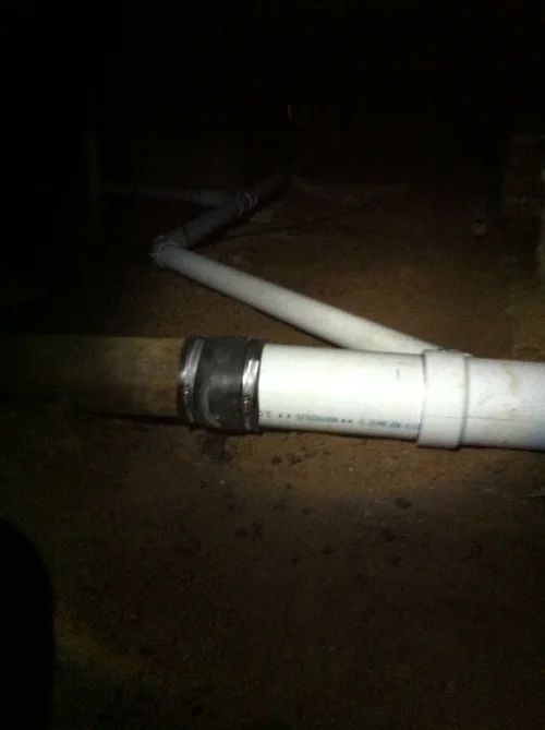 PVC pipe connection in a dark, dirty crawlspace; a coupling joins white and brown pipes.