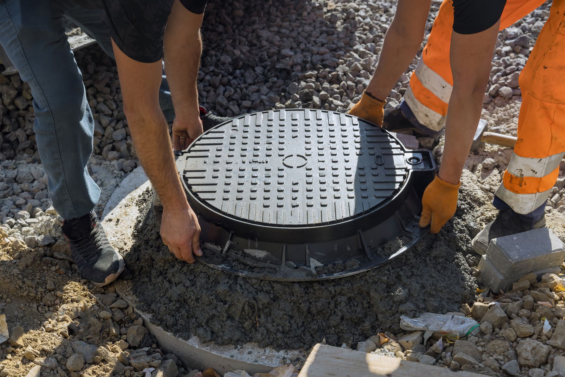 Two workers installing a manhole cover on a concrete base. One wears jeans, the other orange work pants.