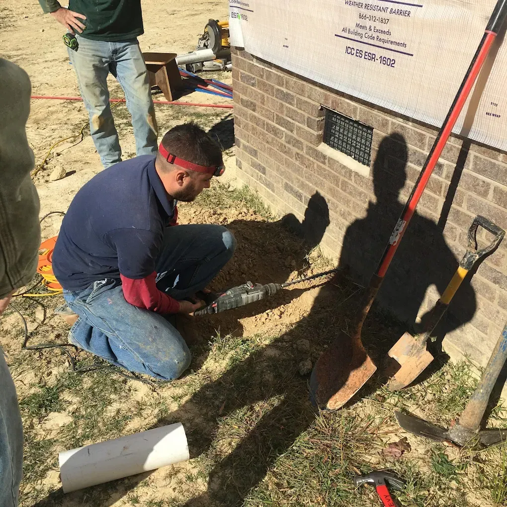 Construction worker kneeling, cutting pipe near a building foundation; a shovel and tools are nearby.