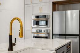 Gold faucet on a white countertop in a modern kitchen with a stainless steel refrigerator and built-in ovens.