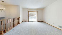 Empty room with light gray carpet, sliding glass door, and chandelier. Staircase railing on the left.