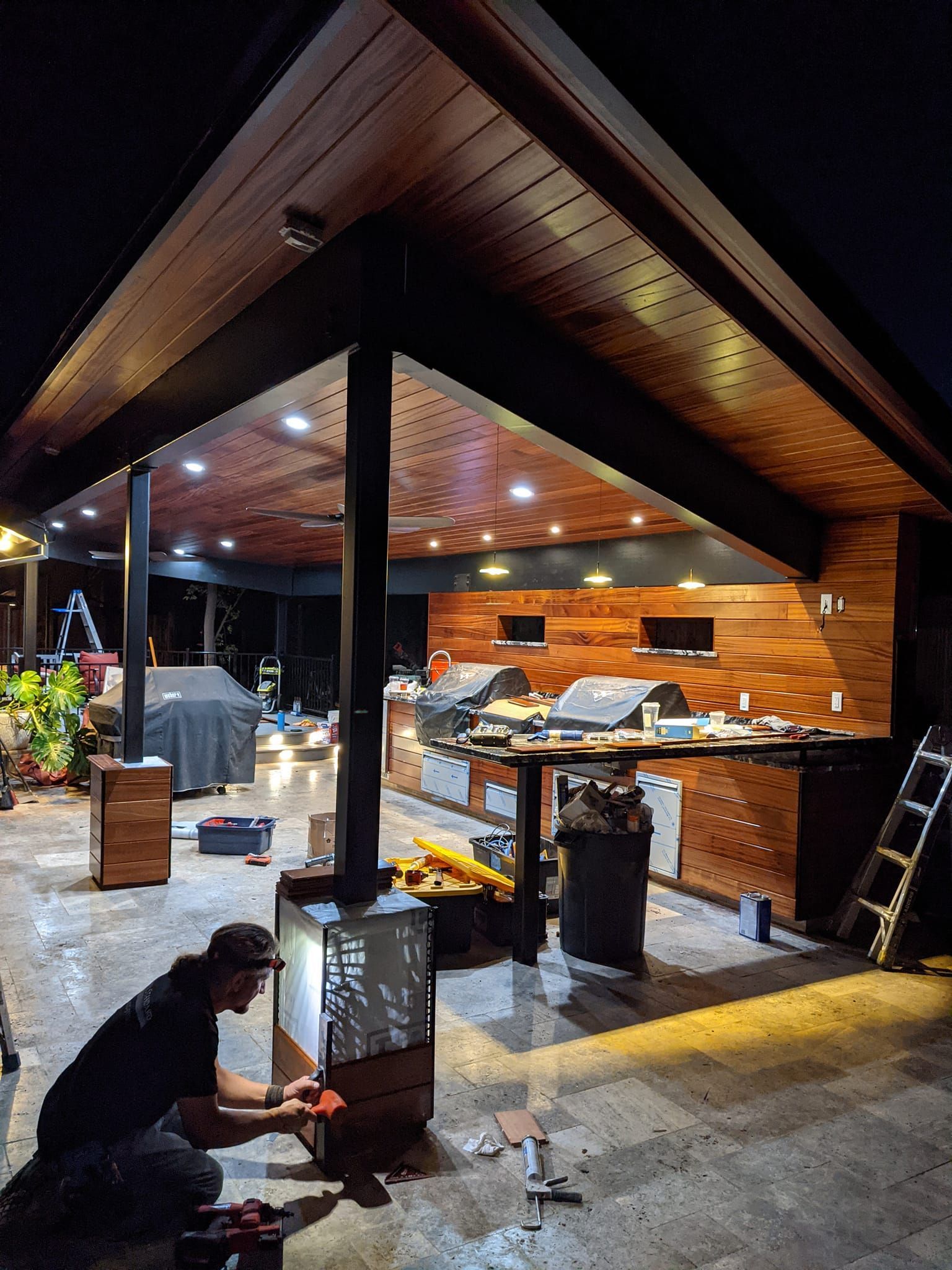 Man working on an outdoor structure with a dark wood ceiling. He is focused, surrounded by tools and equipment at night.