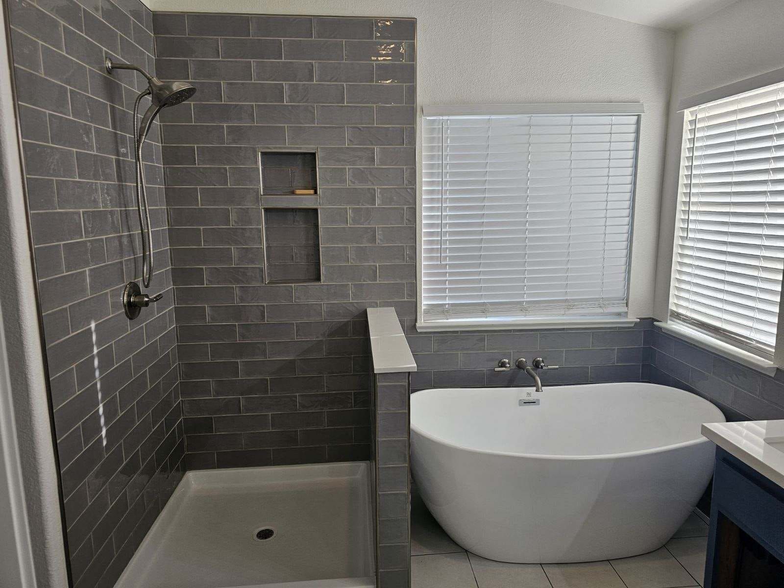 Modern bathroom with gray tiled shower and tub. White soaking tub sits beneath a window with blinds.