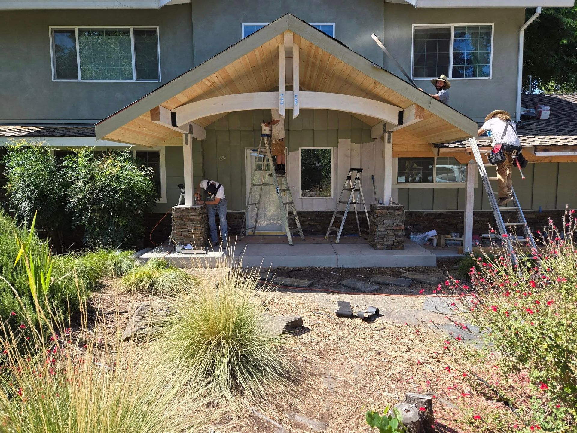 Workers renovating a house's front porch. The porch has stone columns and wooden beams.