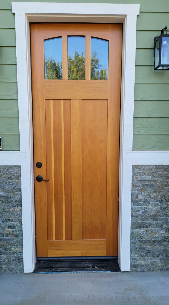 Wooden door with arched glass panels, black hardware, set in white trim against green siding and stone veneer.