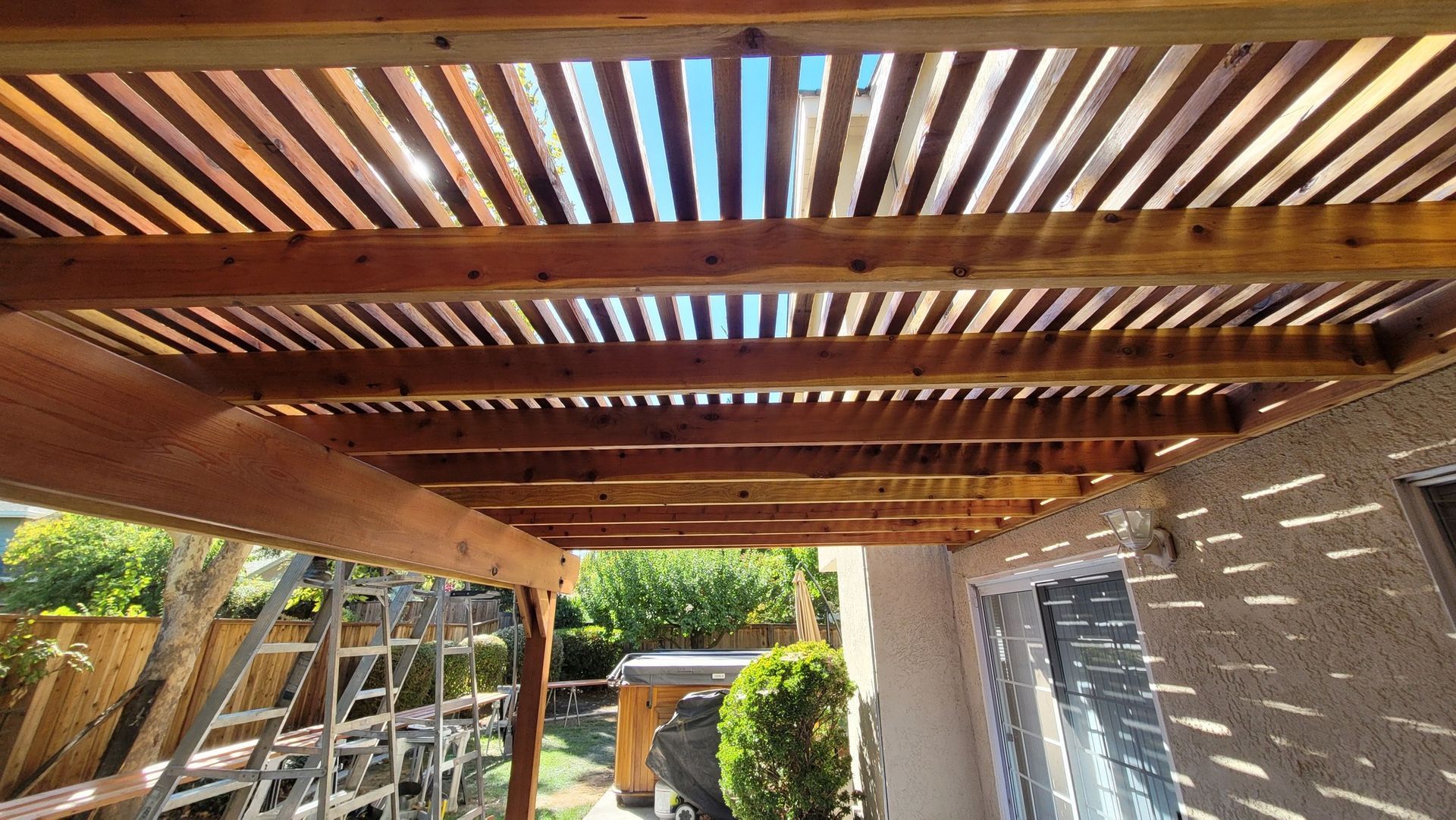 Wooden pergola with slatted roof casting shadows, over a patio area.
