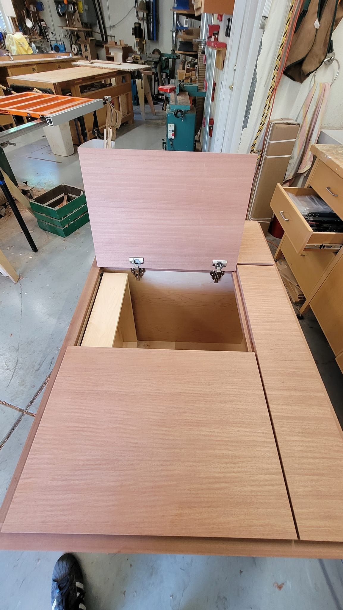 Wooden workshop table with lid open, revealing storage compartment. Other wood and tools in background.