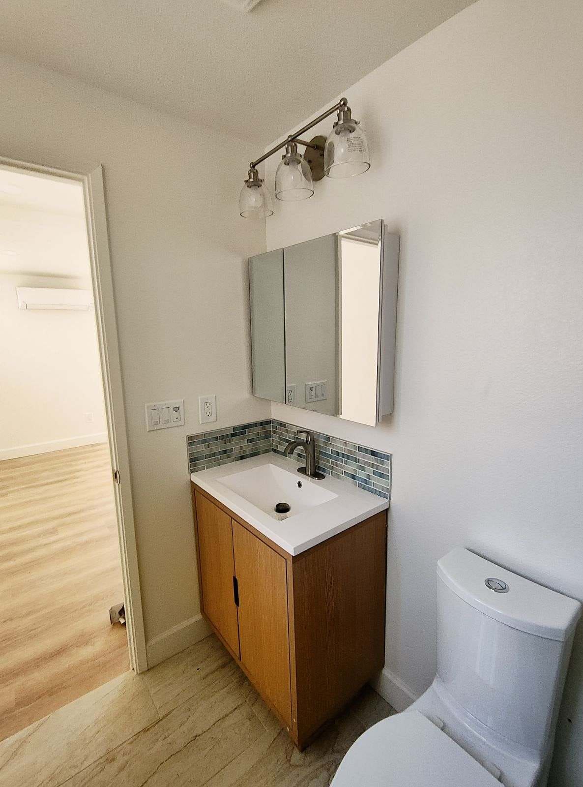 Bathroom with vanity, mirror, toilet, and open door to another room with light wood floors.
