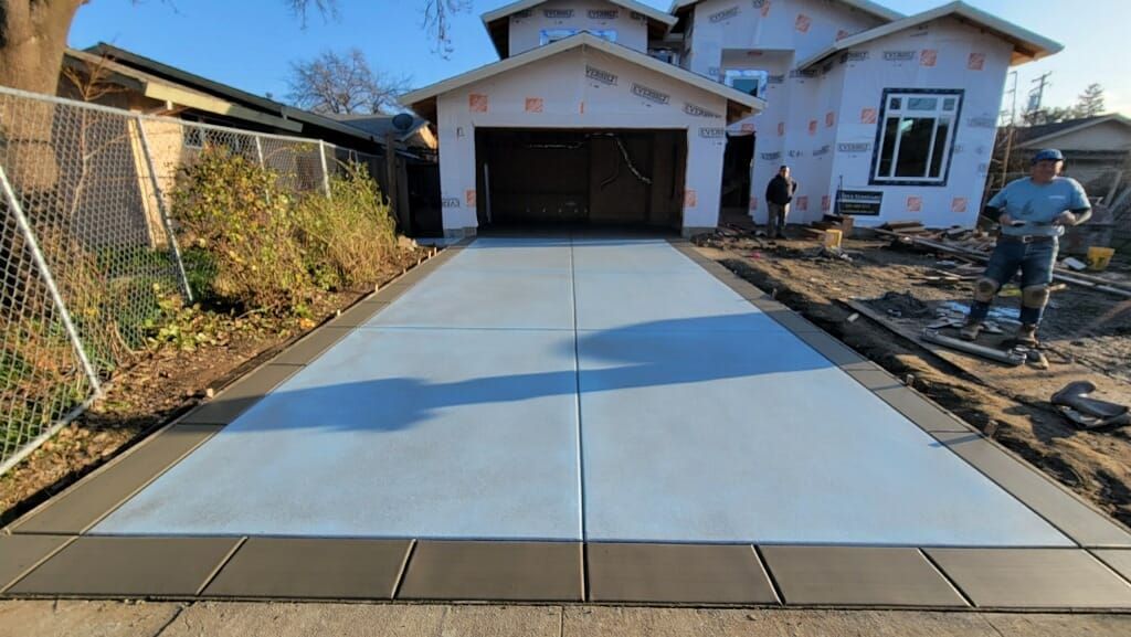 Newly poured concrete driveway with dark border, leading to a garage under construction. A worker stands nearby.