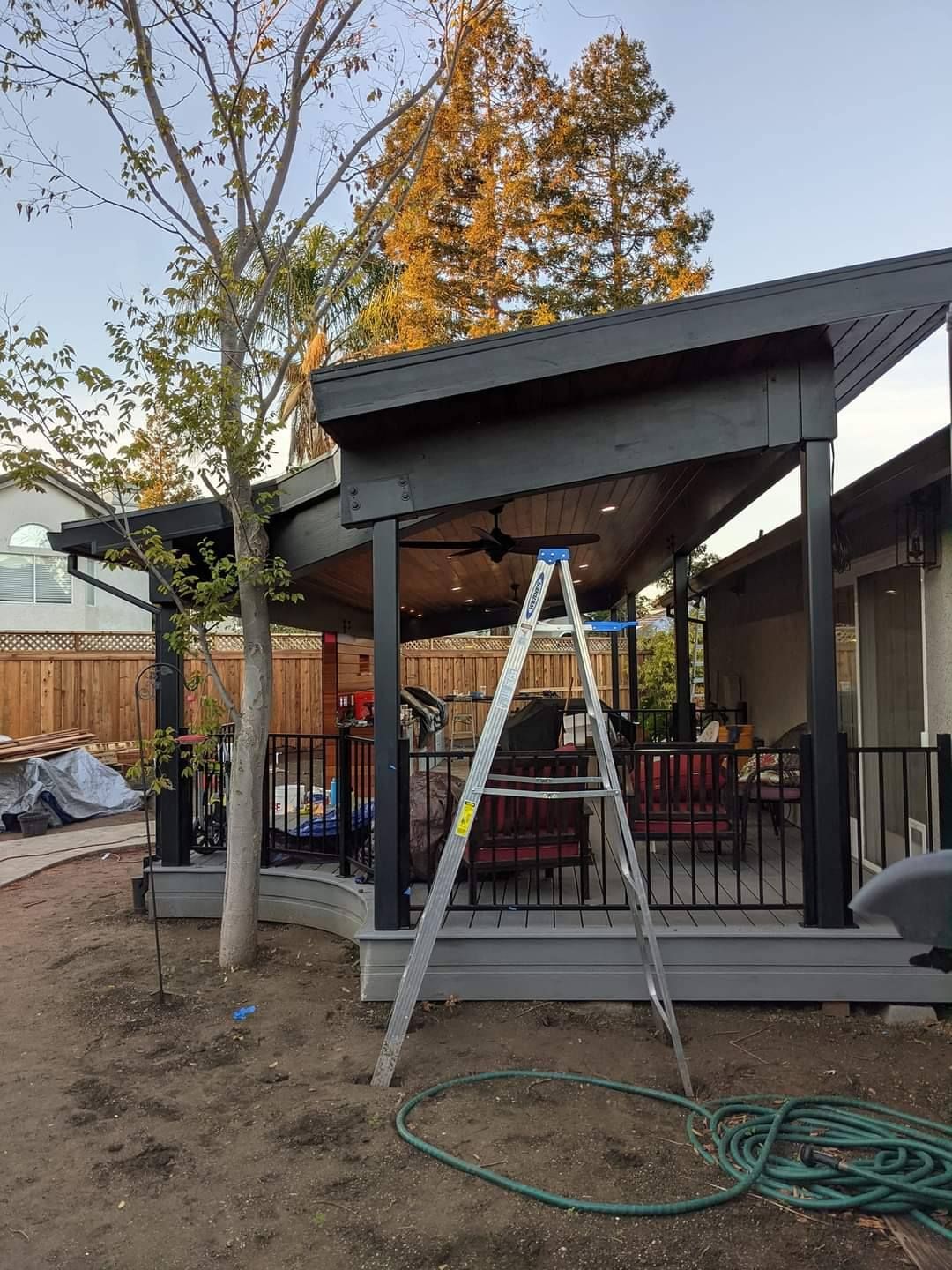 A-frame ladder inside a dark-painted patio cover, overlooking a backyard with furniture and trees.