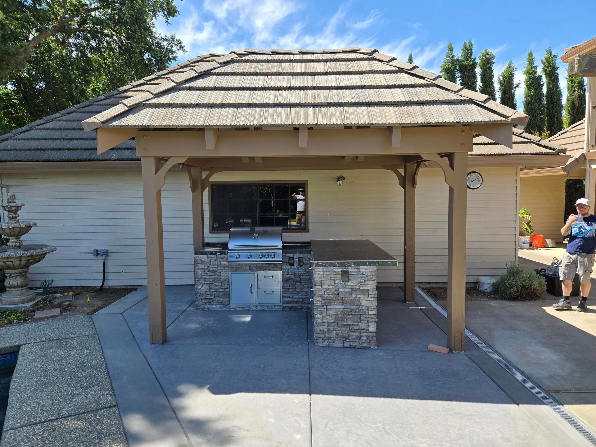 Outdoor kitchen with grill, stone counter, and tiled roof structure. A person stands nearby.