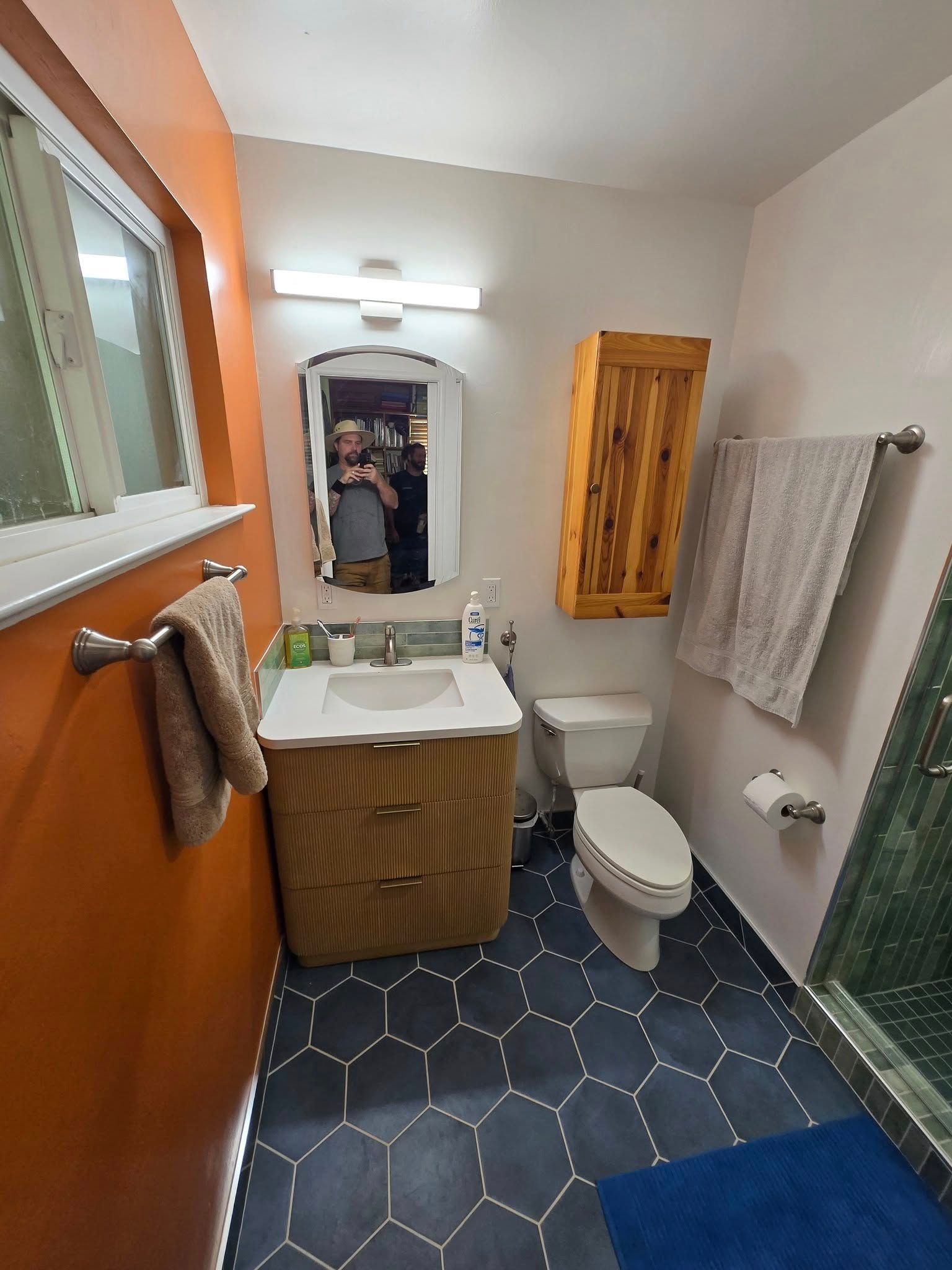 Bathroom with orange and white walls, blue hexagon tile floor, sink, toilet, wooden cabinet and a window.