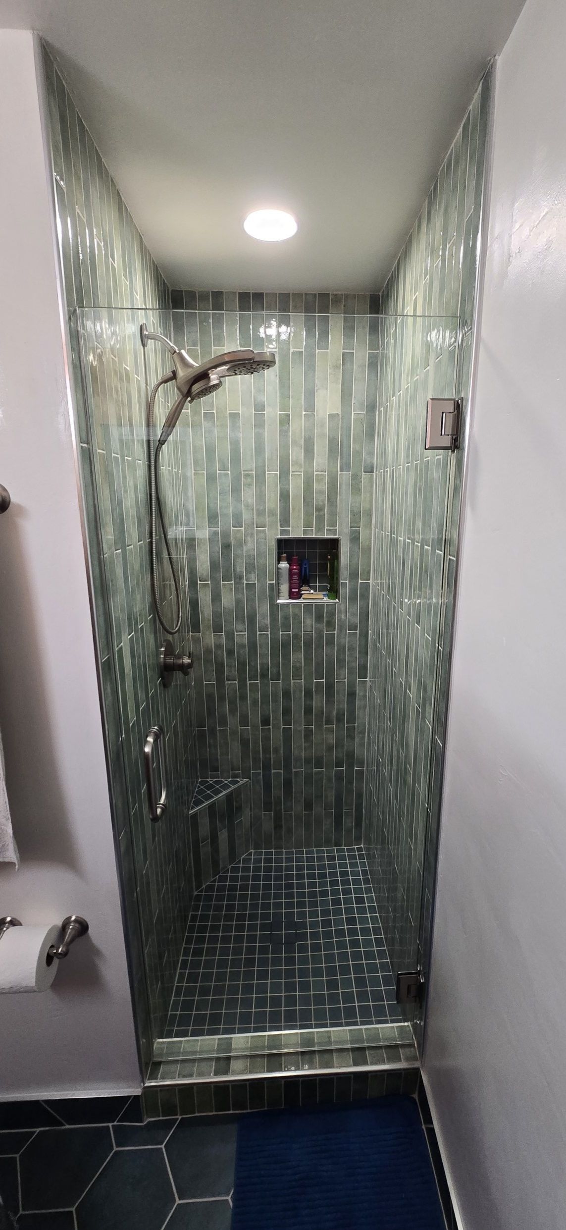 A walk-in shower with green tiled walls and a silver shower head. A blue mat is on the floor.