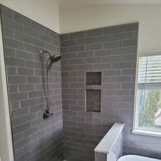 Gray tiled shower with a niche and showerhead, next to a window with blinds.