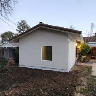 Small stucco building with dark roof, single window, and dirt in the yard.